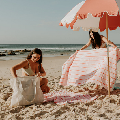 Friends enjoying a day at the beach with their neutral Sande Beach Hauler - Mini beach bags. The scene features pink and red striped beach towels laid out alongside a coral red and white beach umbrella, creating a vibrant and fun atmosphere for a perfect beach day.