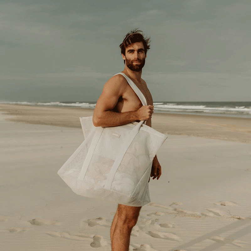 A tall man with brown hair and beard carrying the Sande™ oversized mesh beach bag to the beach. Soft sand with waves int he background.