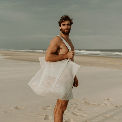 A tall man with brown hair and beard carrying the Sande™ oversized mesh beach bag to the beach. Soft sand with waves int he background.