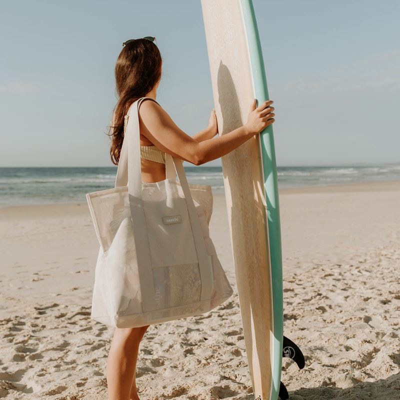 Friends enjoying a day at the beach with their neutral Sande Beach Hauler - Mini beach bags. The scene features pink and red striped beach towels laid out alongside a coral red and white beach umbrella, creating a vibrant and fun atmosphere for a perfect beach day.