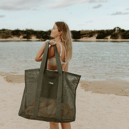 A women holding the Sande™ Beach Hauler oversized mesh beach bag in Pandanus green at a lake beach with dune sin the background.