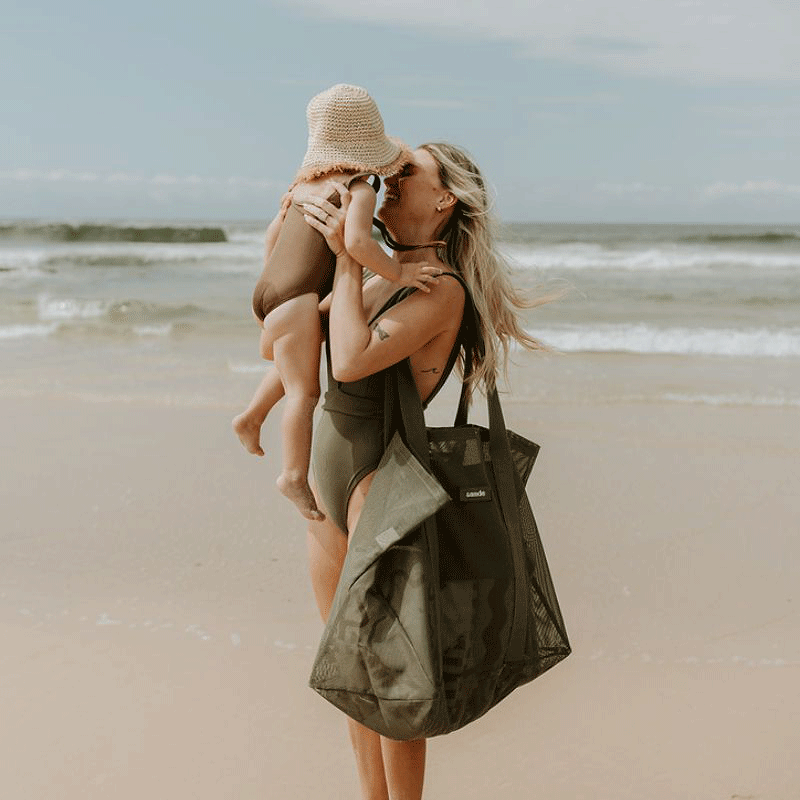 Woman at the beach holding her toddler up to her face, carrying the Sande Kids Beach Hauler oversized mesh beach bag in Pandanus on her shoulder, filled with beach essentials.