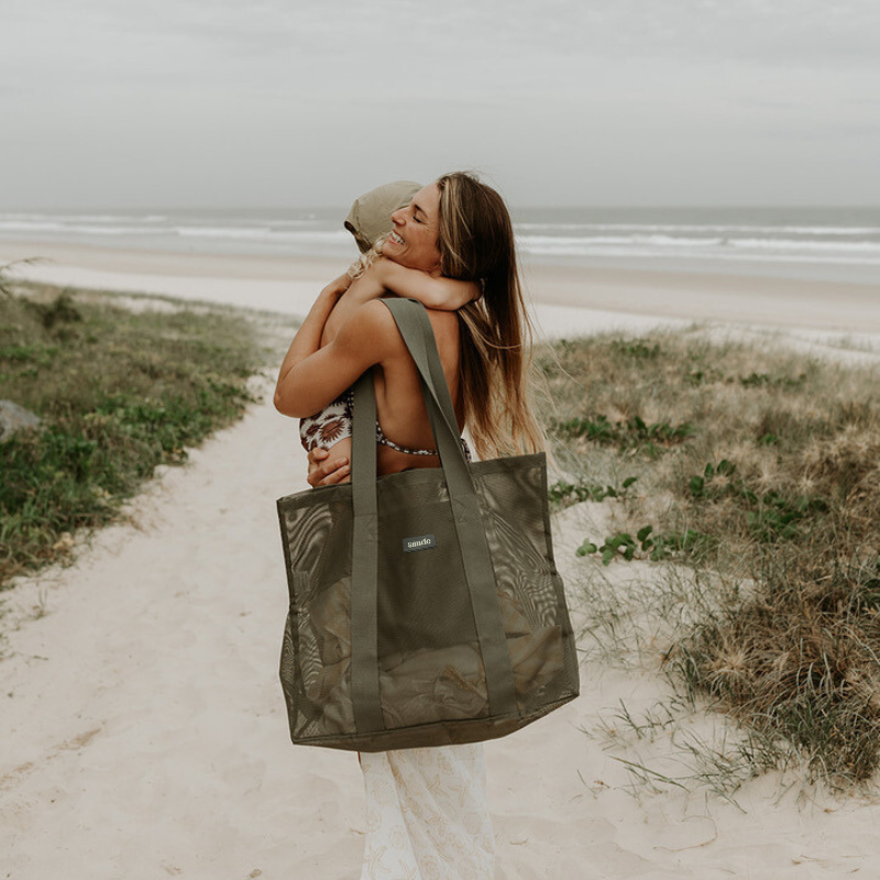 Mother at the beach carrying her toddler son, with a Sande Beach Hauler Mini in Pandanus green on her shoulder, showcasing its convenient size and family-friendly design for beach outings.