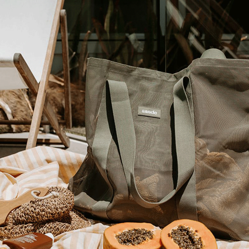 Oversized mesh beach bag in olive green beside a swimming pool, with a striped beach blanket, and a white canvas beach chair with wooden frame in the background.