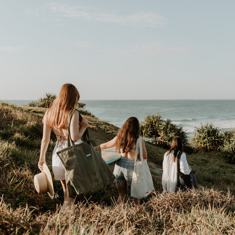 A group of girlfriends walking down a grassy headland at Cabarita Beach, carrying surfboards and their Sande Beach Hauler - Mini beach bags. This scene captures the essence of fun and friendship at the beach, showcasing the stylish and functional design of the Sande Kids bags.
