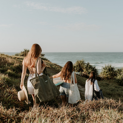 A group of girlfriends walking down a grassy headland at Cabarita Beach, carrying surfboards and their Sande Beach Hauler - Mini beach bags. This scene captures the essence of fun and friendship at the beach, showcasing the stylish and functional design of the Sande Kids bags.
