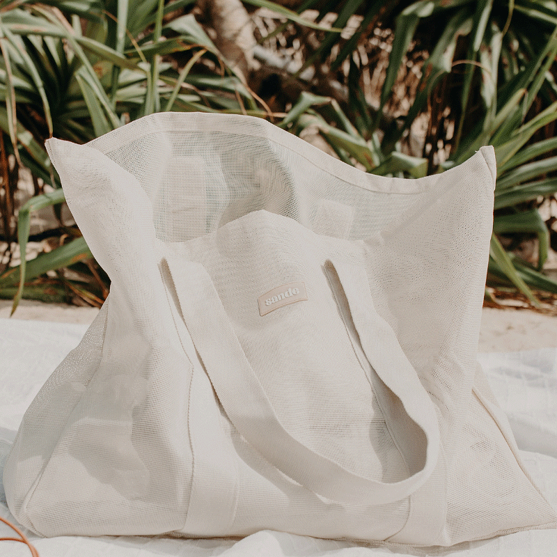 Sande™ Beach Hauler oversized PVC-coated polyester mesh beach bag in Sand, displayed on a white beach blanket on the sand with pandanus palms in the background.