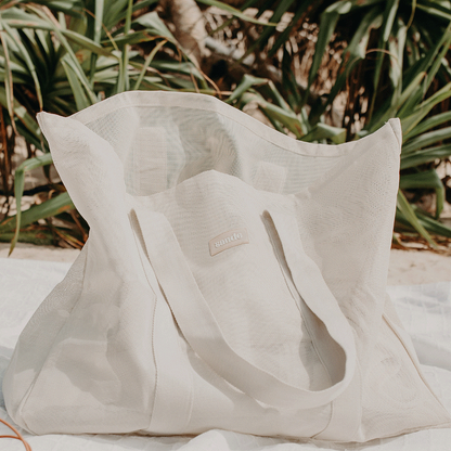 Sande™ Beach Hauler oversized PVC-coated polyester mesh beach bag in Sand, displayed on a white beach blanket on the sand with pandanus palms in the background.