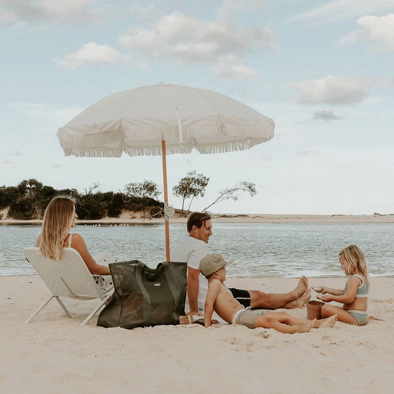 A young family hanging a a lake beach. White Basil Bangs and beach chair, family lazing arounds. A Sande Kids Beach Hauler mesh beacg bag in Pandanus feint and centre.