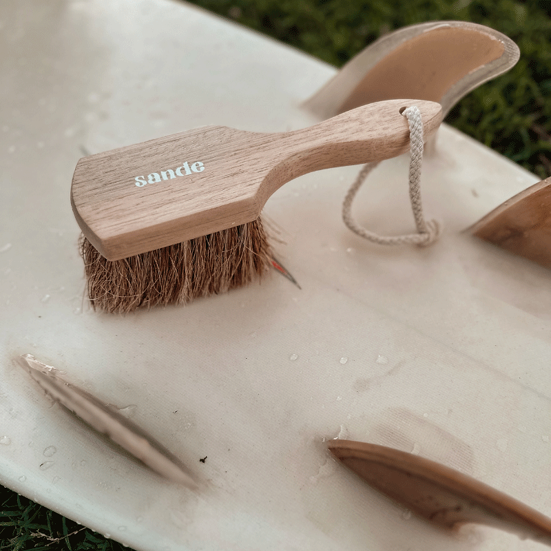 Sande natural sand remover brush sitting on a surfboard with natural colour surfboard fins.