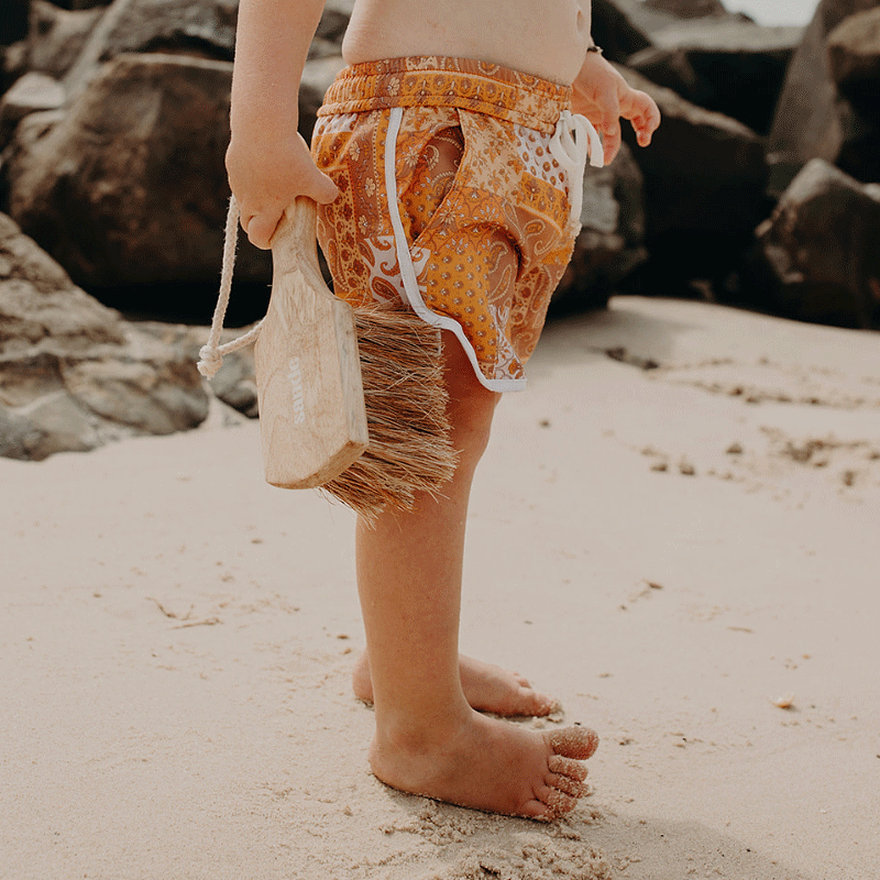 A little boy standing on the beach, holding the Sande™ Sand Brush. Wearing orange board shorts, he is ready to sweep sand off his feet, with black rocks in the background.