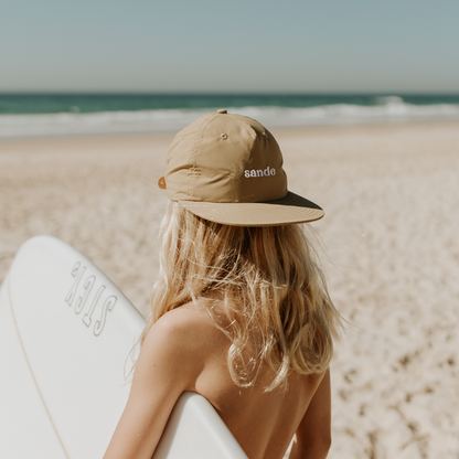 Lifestyle image of a young surfer with long blonde hair, holding a surfboard under one arm on the beach, looking at the ocean, wearing a beige quick-dry Sande cap backwards.