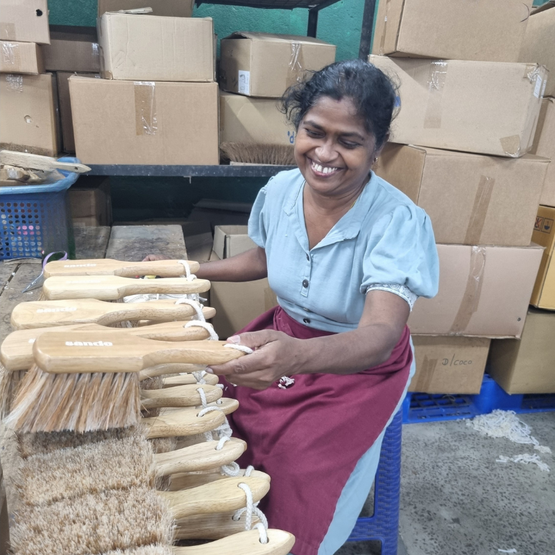 A smiling woman sorting the Sande sand brush in the factory in Sri Lanka. She is sitting on a blue stool with sand brushes in front of her. There are packing boxes in the background.
