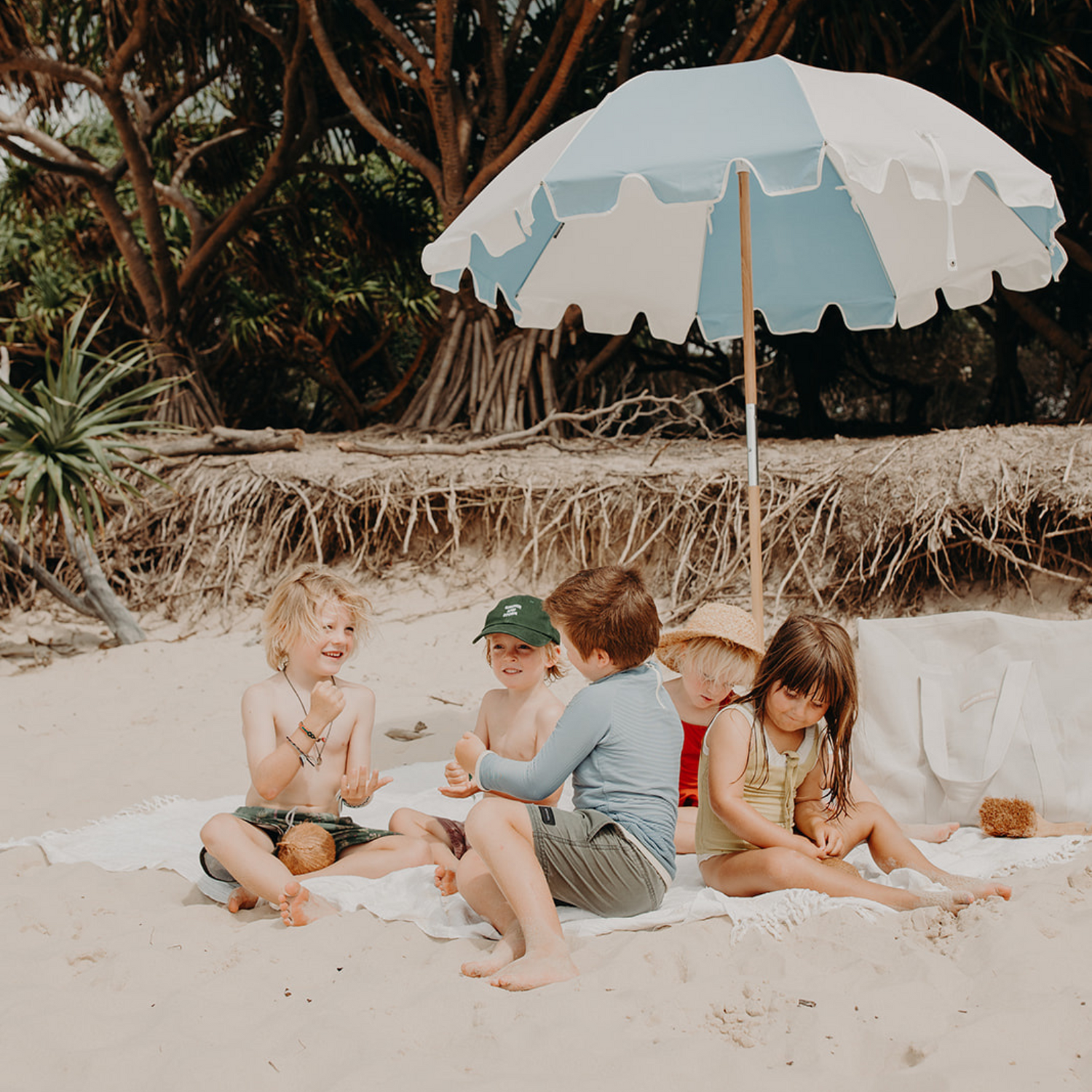 Five young children sitting on a white beach blanked under a blue and white panelled beach umbrella. Pandanus palm trees in the background.